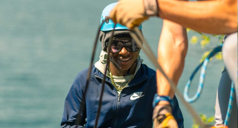 In the foreground, a two hands manage a rope, while a person wearing rock climbing gear stands behind them on a cliff above blue water 